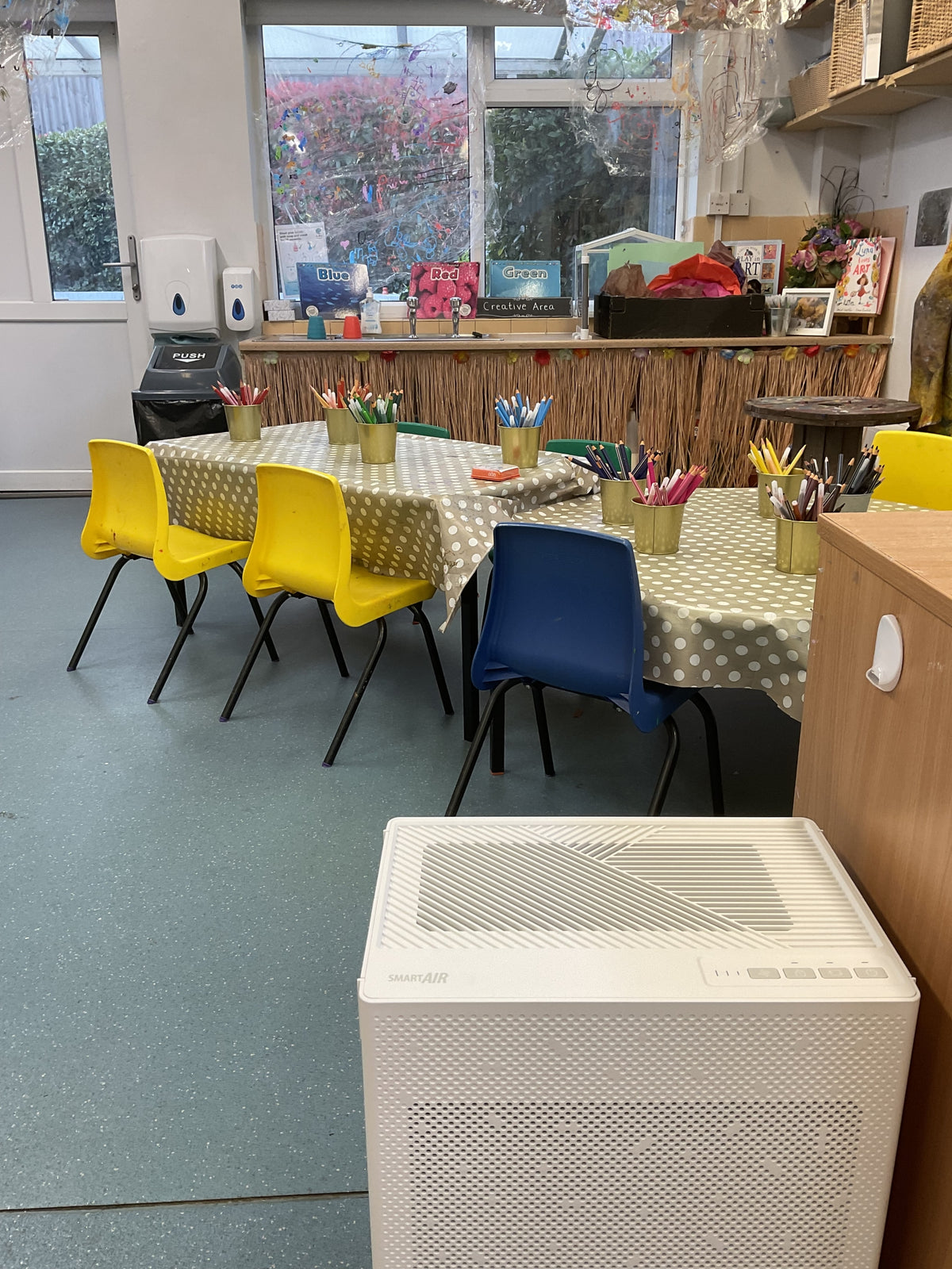 A white portable air purifier with a perforated front grille sits on a gray floor in a classroom or playroom setting with children's tables, colorful chairs, and art supplies in the background.